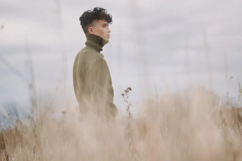 A solitary figure stands in tall grass wearing a chunky turtleneck, conveying authenticity and credibility through a quiet gaze and natural, muted light.