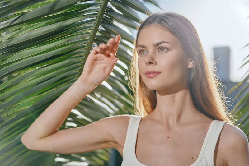 Young woman standing outdoors near tropical palm leaves with sunlight on her face, wearing a white tank top, looking serene and thoughtful.