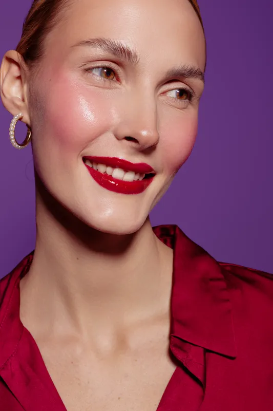 Close-up of a smiling woman with glossy red lipstick and radiant skin wearing a red blouse and hoop earrings on purple background. Concept of beauty, makeup, and skincare.