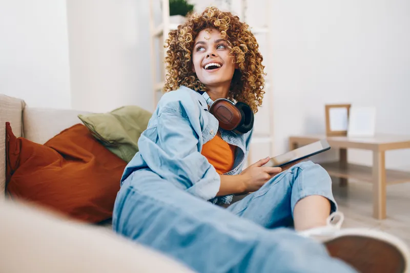 Улыбающаяся женщина держит планшетный компьютер и слушает музыку в наушниках, сидя на диване дома