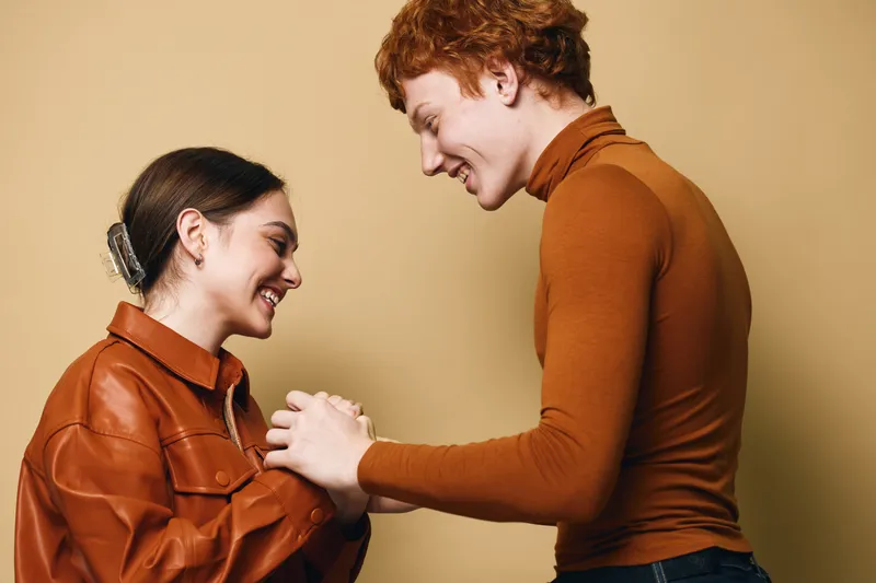 Young couple holding hands and smiling, facing each other with affection against beige background. Casual clothing with warm colors and genuine emotion between two people.