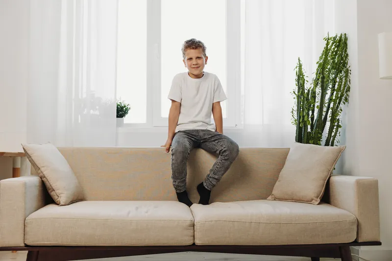 A smiling boy in casual clothes sits on a beige sofa in a bright living room with white curtains and green plants. Concept of childhood, home, and comfort.