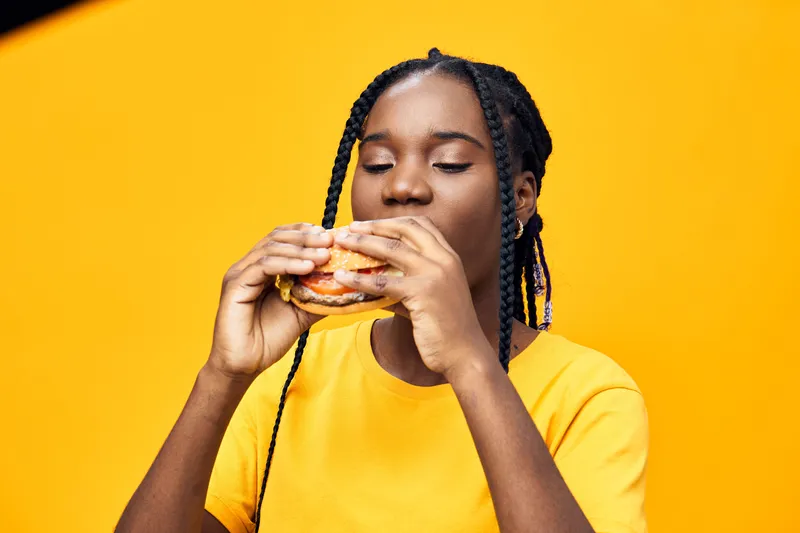Young woman enjoying a delicious burger against a vibrant yellow background, showcasing her joyful expression, ideal for promoting healthy eating or fast food concepts