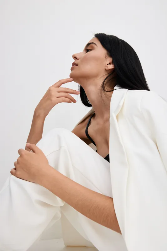 Young woman in white suit poses gracefully against plain background, showcasing modern fashion style with calm, confident expression in minimalistic setting.