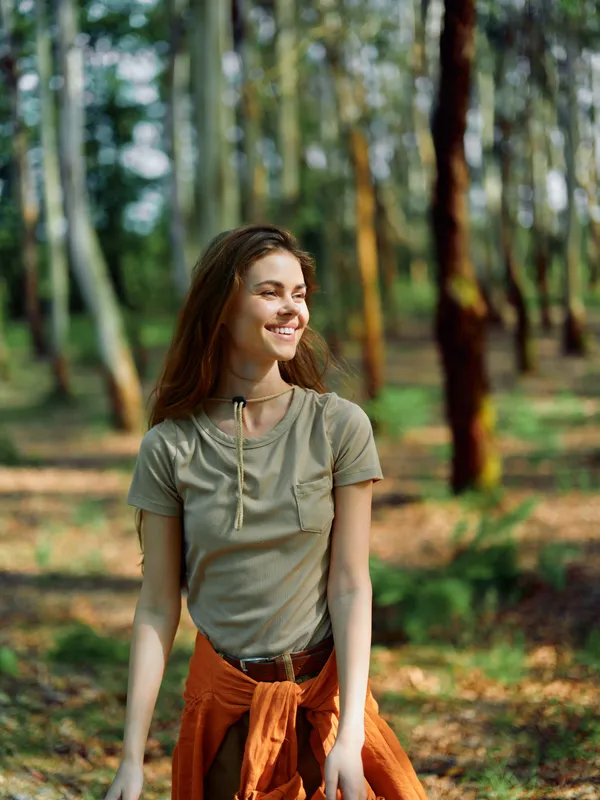 Woman smile forest nature sunlight portrait outdoors walking, young woman smiles in sunlit woodland wearing casual clothes with tied jacket, relaxed and happy enjoying peaceful nature.