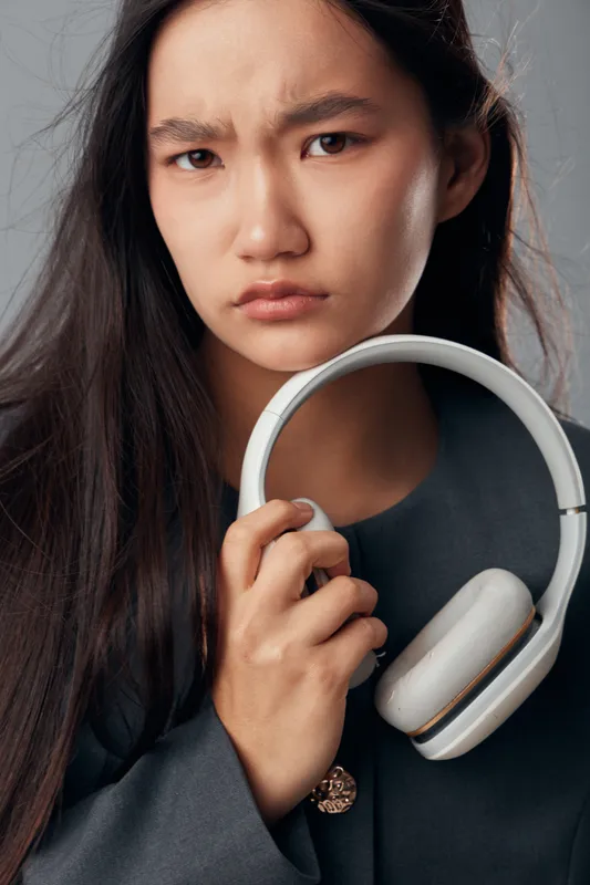 Young woman holding headphones with serious facial expression closeup portrait on neutral background. Female with long dark hair showing concern or dissatisfaction indoors.