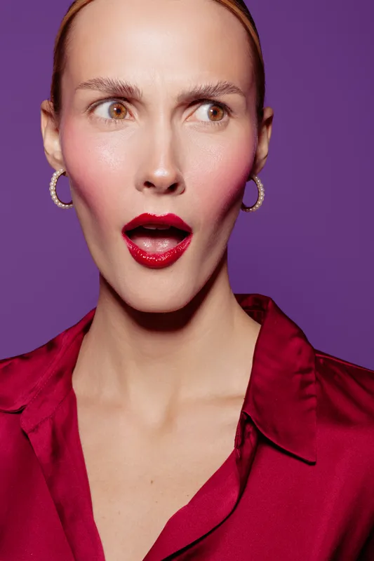 Surprised woman with red lipstick and blush looks sideways on purple background. Fashion portrait with expressive face and elegant red blouse. Concept of beauty and emotion.