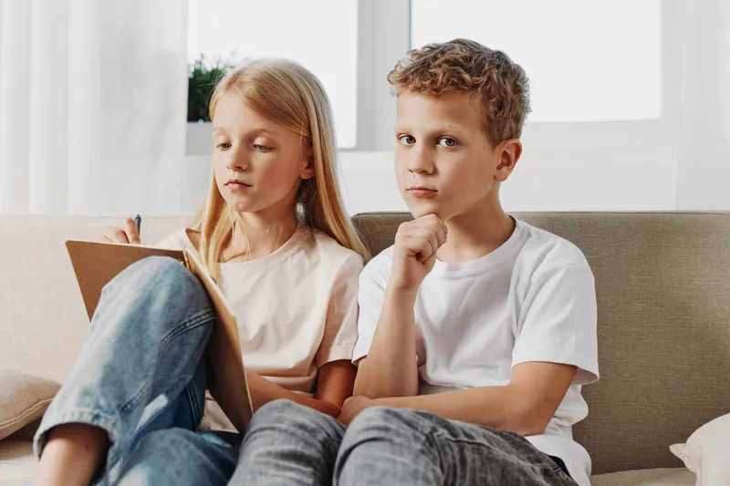Thoughtful boy and focused girl sit on couch in casual clothes, boy resting chin on hand while girl writes on clipboard. Concept for concentration, childhood, and learning at home.