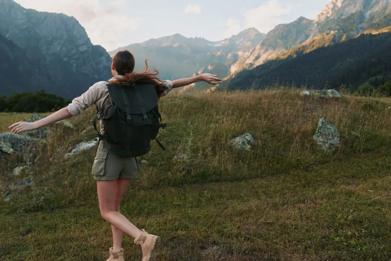 Joyful woman outdoors, arms raised, hiking traveler enjoying vast mountains and valley, sunlit meadow, natural colors, serene moment, active lifestyle, bright weather, sense of freedom