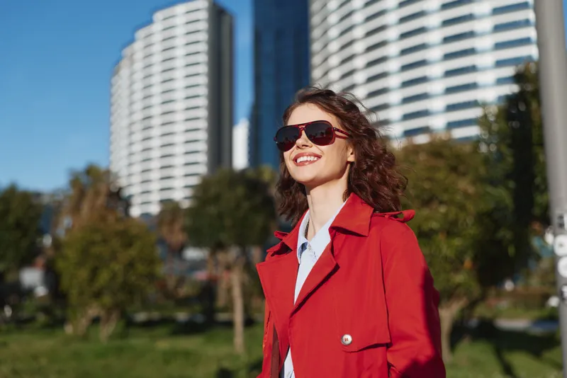 A young woman wearing a red coat smiles confidently in a sunny city park with tall modern buildings in the background, conveying casual street style and upbeat energy.