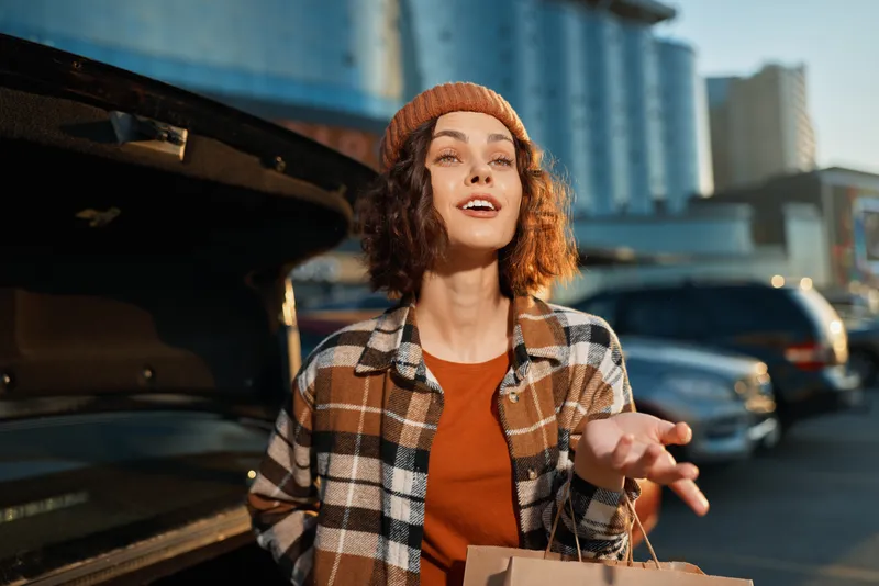 Woman portrait by car in urban parking, golden sunlight and candid lifestyle moment with shopping bag, beanie and plaid jacket conveying authenticity, mindful living and emotional storytelling.