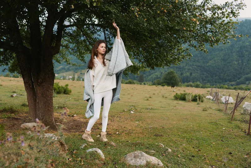A woman stands in a grassy field beside a large tree, wearing a light outfit and a draped blanket, posing for a lifestyle photo about calm outdoor living.