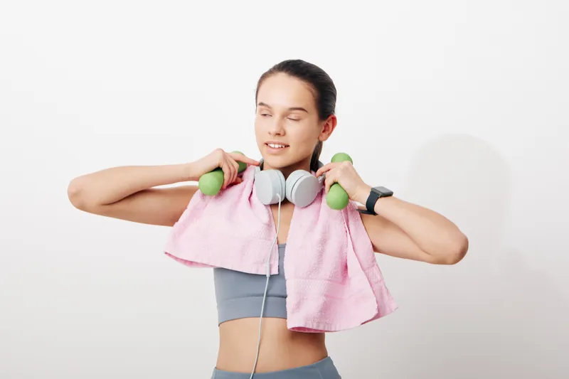 Woman in athletic wear lifts light dumbbells with a pink towel draped over her shoulders, showcasing a focused workout routine, fitness dedication, and active lifestyle in a clean studio.