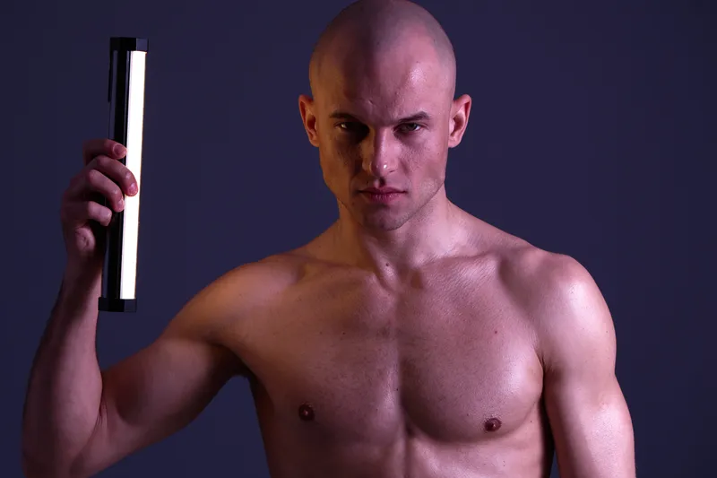 A shirtless man holding a light stick confidently, showcasing strength and intensity in a dramatic black and white composition, emphasizing emotion and determination.
