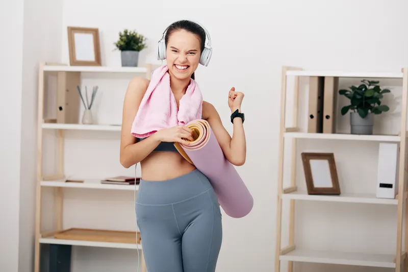 Woman fitness enthusiast with headphones, holding a rolled yoga mat and towel, posing in a bright room with bookshelves and plants, ready for workout, training, and athletic performance.