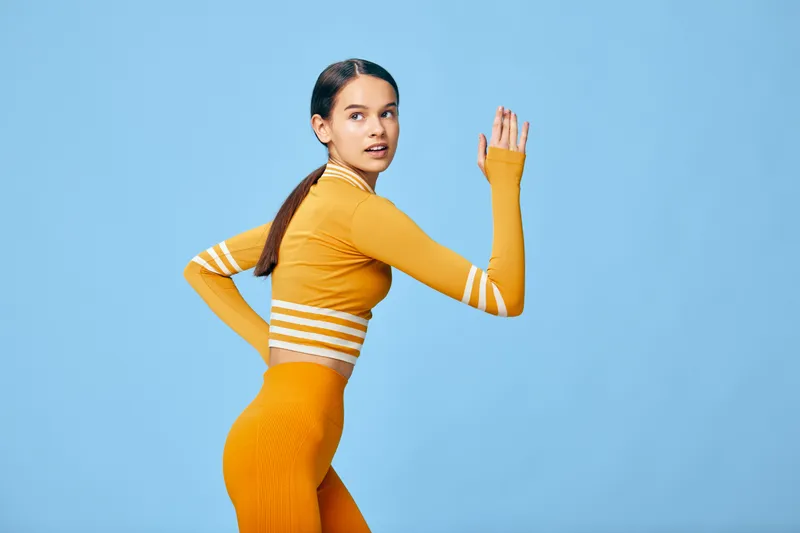 Young woman in orange fitness outfit posing in a running stance against a solid light blue studio background, crop striped top, high waist leggings, ponytail, athletic posture.