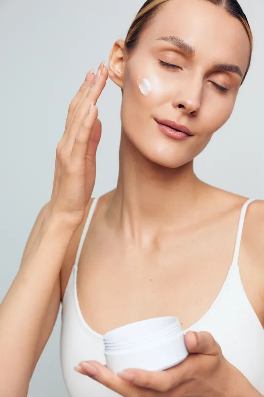 A young woman in a white tank top applies cream to her cheek with closed eyes. The clean background highlights skincare and beauty concept with natural skin and gentle care.