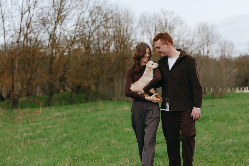 Cheerful couple outdoors, smiling as they cradle a goat kid during a relaxed park stroll, sunlit scene, gentle breeze, natural colors, candid lifestyle moment of warmth and connection