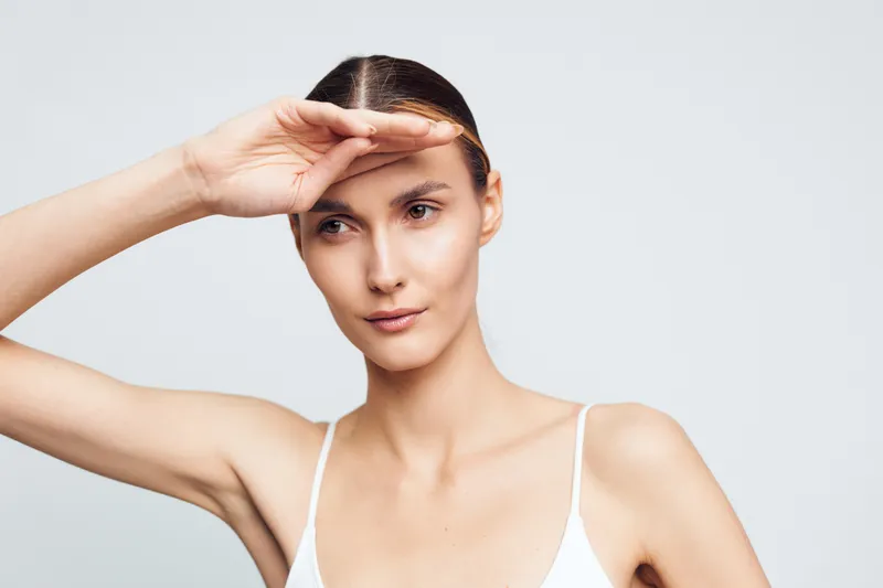 Young woman with dark hair shields her eyes with her hand in a bright studio, conveying focus and subtle curiosity in a bright studio with soft lighting and a minimal backdrop for lifestyle