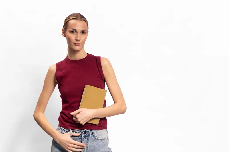 Young woman with blonde hair in red sleeveless top holds a brown notebook against white background. Casual style, confident pose, concept of education and lifestyle.