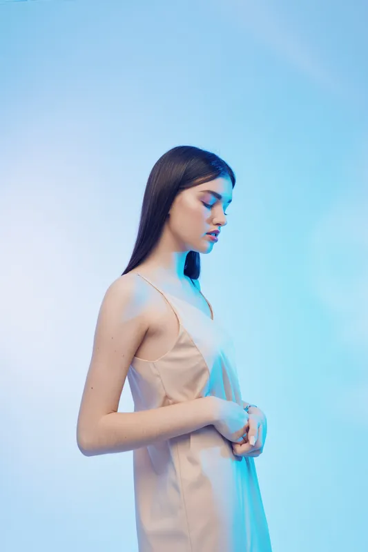 Young woman in beige dress poses with eyes closed against blue background. Soft lighting highlights her smooth skin and elegant beauty. Concept of fashion and beauty.