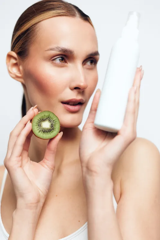 A woman holds a kiwi beside a white moisturizer bottle, illustrating natural skincare, hydration, and a healthy complexion in a fresh, minimal beauty routine.