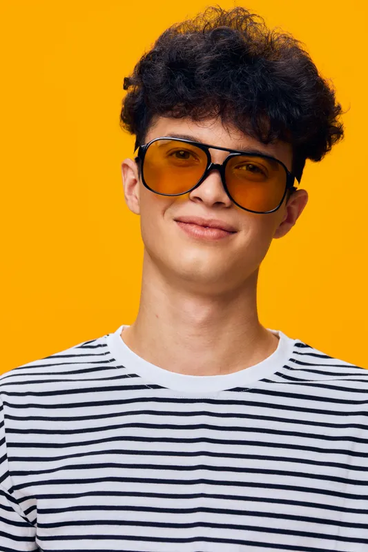 young man wearing yellow sunglasses and striped shirt smiling against vibrant orange background, curly hair, casual and stylish appearance