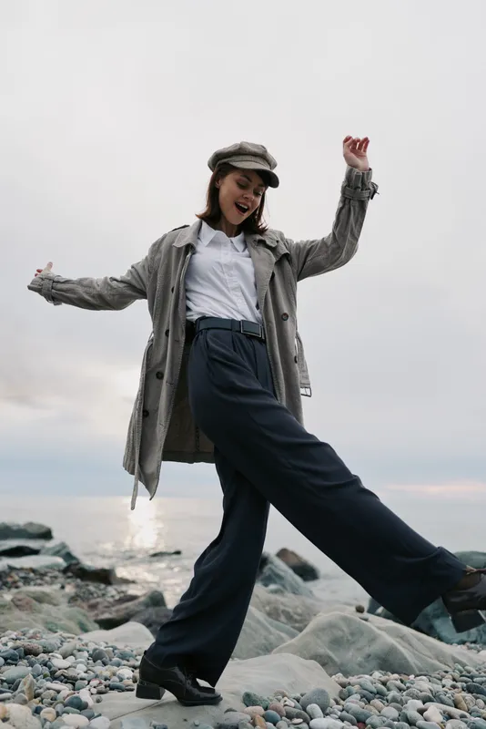 Young woman in stylish coat and hat enjoys outdoors on rocky beach, smiling and lifting leg. Casual fashion and joyful mood captured during cloudy day near water.