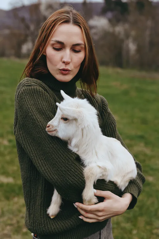 Cheerful woman outdoors, cradling a white goat kid in a knitted sweater, soft smile, sunlit grass, gentle breeze, natural colors, tranquil setting, warm, intimate moment, rustic charm