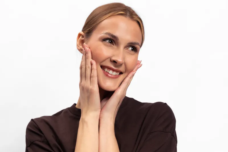 Smiling young woman with clear skin touches her face gently, looking to the side. Studio portrait on white background. Concept for skincare, beauty, wellness, and self-care.