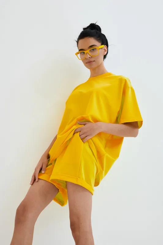 young woman in yellow outfit wearing stylish glasses posing confidently against white background, casual summer attire, modern fashion look