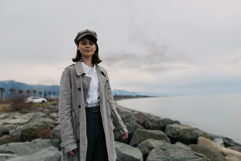 Young woman in a plaid coat and cap stands on rocky shore near calm sea under cloudy sky. Casual autumn outfit, natural light, outdoor lifestyle and serene coastal mood.