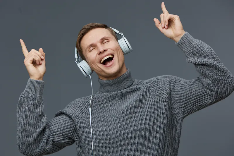 Joyful man with headphones expressing happiness, dancing and enjoying music in a casual gray sweater, against a plain gray background. People lifestyle concept, positive emotion, leisure activity, rel