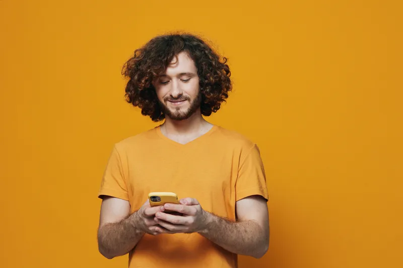 Young man with curly hair in a yellow t shirt using a smartphone against a vibrant orange background, conveying joy and connection through technology