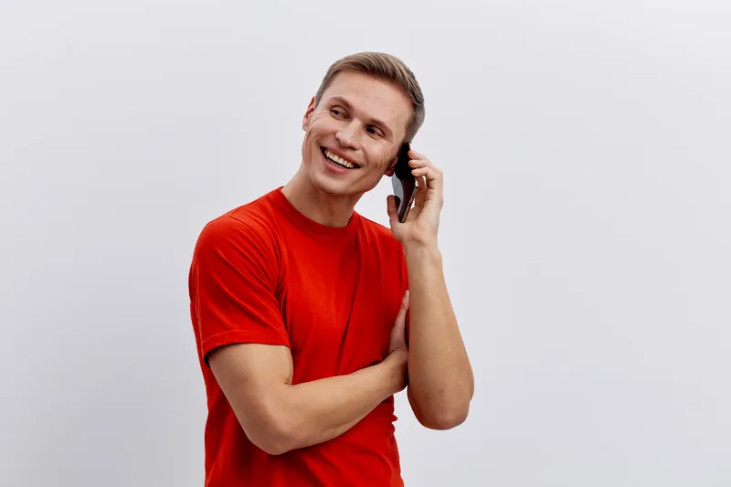 Smiling man with short blond hair wearing a bright red T-shirt, talking on mobile phone, friendly expression, casual style, neutral background, studio shot, lifestyle concept, cheerful mood