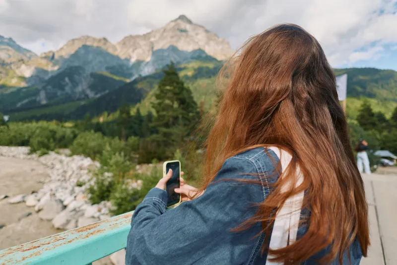 A person with long hair stands on a mountain trail, using a smartphone to capture the scenery. The panoramic landscape features rocky peaks, lush trees, and bright daylight.
