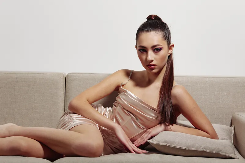 Young woman with sleek long hair poses confidently on beige sofa in minimal satin dress. Bold makeup and creative lighting emphasize beauty and fashion styling for modern editorial concept.