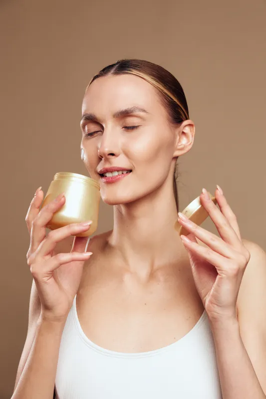 Woman with smooth skin applying cream from small jars closeup beauty skincare concept wellness selfcare moment calm expression soft studio lighting on brown background