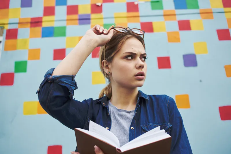 Студентка в рубашке и очках держит в руках книгу на улице возле граффити на стене