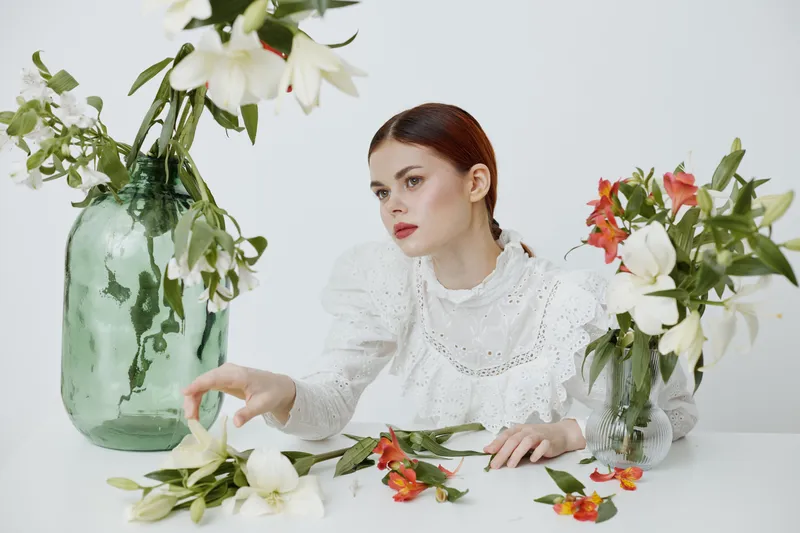 A serene young woman in a white lace blouse arranging fresh flowers in green vases, conveying tranquility and beauty in a minimalist setting