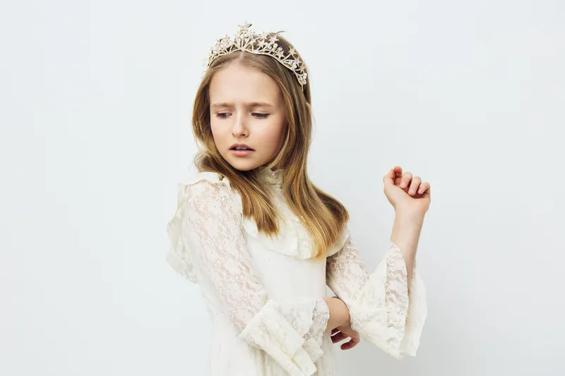 Portrait of a young girl in a delicate lace dress and tiara, expressing elegance and innocence against a soft white background