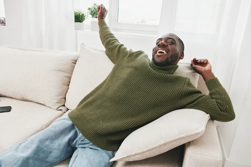 Cheerful young African man in a cozy green sweater smiling and relaxing on a light sofa at home, depicting comfort and leisure