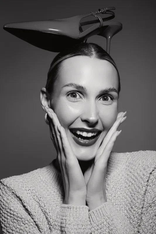 Happy Caucasian woman with light skin smiles widely, hands on cheeks, high heel shoe balanced on head. Studio portrait with dramatic lighting.