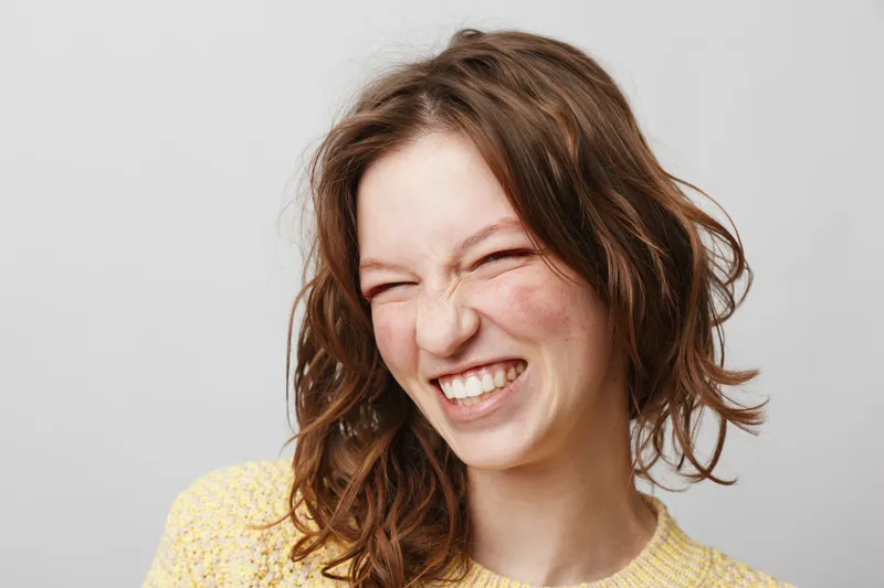 Young woman with curly hair smiles with eyes closed wearing yellow sweater. Studio portrait on light background. Concept of happiness, joy, and natural beauty.
