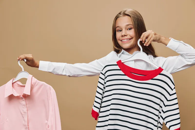 Girl smiling and holding two clothes hangers with different shirts for choosing outfit in casual style at home or studio with beige background.
