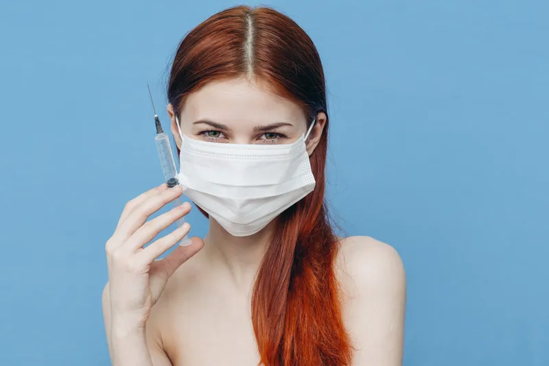 Женщина на голубом фоне в медицинской маске, в руке шприцы — укол ботокса, инъекция красоты, уход