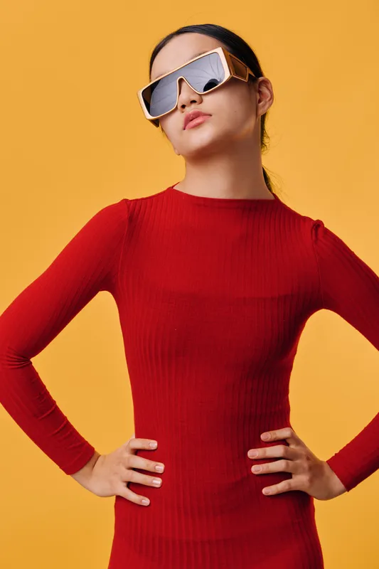 Woman in red dress wearing modern sunglasses posing confidently with hands on hips against yellow background. Fashion, style, youth and attitude with trendy eyewear and casual look.