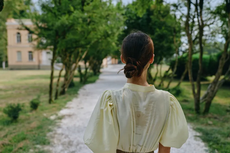 woman back walking on park path with vintage dress and trees, authentic inclusive portrait expressing diversity and calm, outdoor nature greenery and historic building in soft light