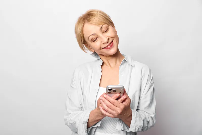 Senior woman uses smartphone with relaxed smile, showing technology and messaging for social media communication, indoor studio portrait against gray background, holding device in white shirt,