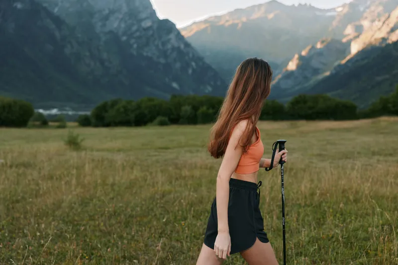 A young woman hikes through a grassy valley with towering mountains in the background. She wears a coral top and dark shorts, using trekking poles on a sunny day.